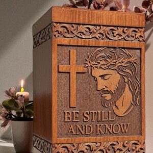 Close-up view of the “Be Still and Know” wooden cremation urn box showing intricate engraving of Jesus’ face and the cross. The fine detailing and smooth craftsmanship highlight premium wood texture and artisan engraving. A spiritual and meaningful Christian funeral urn representing comfort, belief, and everlasting memory.