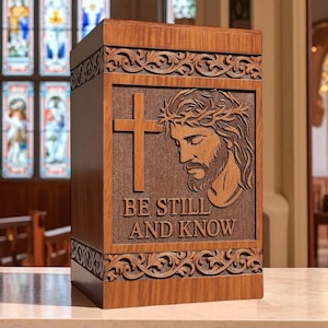 Handcrafted wooden cremation urn box featuring an engraved image of Jesus with a cross and the words “Be Still and Know.” This elegant Christian memorial urn is made from solid wood with intricate floral carved borders, polished smooth for a timeless finish. Ideal for adult human ashes, it serves as a personalized religious keepsake symbolizing peace, faith, and eternal remembrance. Custom engraving available.
