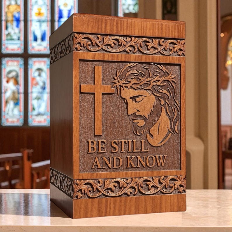Handcrafted wooden cremation urn box featuring an engraved image of Jesus with a cross and the words “Be Still and Know.” This elegant Christian memorial urn is made from solid wood with intricate floral carved borders, polished smooth for a timeless finish. Ideal for adult human ashes, it serves as a personalized religious keepsake symbolizing peace, faith, and eternal remembrance. Custom engraving available.