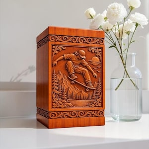 May include: A rectangular wooden urn featuring a detailed relief carving of a person skateboarding over a landscape with mountains, pine trees, and a sun. The carving is framed by decorative scrollwork borders on the top and bottom edges.