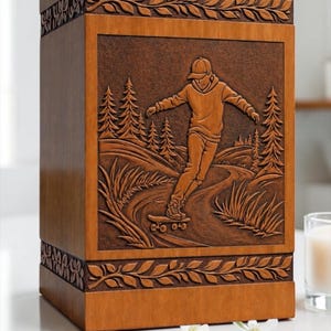 May include: A rectangular wooden cremation urn featuring a carved relief panel on the front. The carving depicts a person wearing a hooded sweatshirt and cap, riding a skateboard on a path through a forest with pine trees and tall grasses.