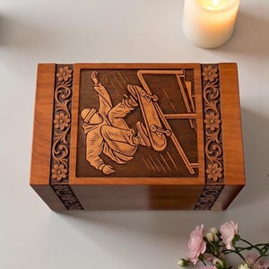 Decorative wooden urn box with skateboard engraving, handmade craftsmanship, carved floral patterns, rich brown polish, secure lid, and sympathy gift display with candle and flowers.