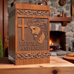 Wooden “Be Still and Know” urn box displayed on a table with candles and flowers, creating a peaceful memorial setting. The engraved image of Jesus with the cross adds divine serenity. This handcrafted Christian cremation urn honors your loved one with dignity, beauty, and faith-inspired remembrance.