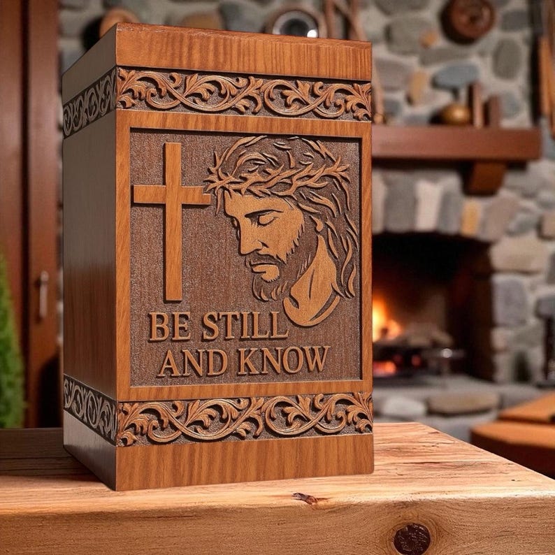 Wooden “Be Still and Know” urn box displayed on a table with candles and flowers, creating a peaceful memorial setting. The engraved image of Jesus with the cross adds divine serenity. This handcrafted Christian cremation urn honors your loved one with dignity, beauty, and faith-inspired remembrance.