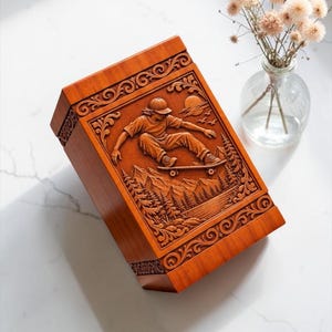 May include: A rectangular wooden box featuring a detailed relief carving on the lid. The carving depicts a person skateboarding over a landscape of mountains, pine trees, and a lake, framed by decorative scrollwork and a sun in the sky.