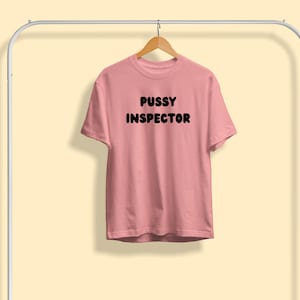 Pu&ograve; includere: Una maglietta rosa chiaro con la scritta "PUSSY INSPECTOR" stampata in grassetto nero. La maglietta &egrave; appesa a una gruccia di legno sospesa a un appendiabiti di metallo bianco su uno sfondo giallo chiaro.