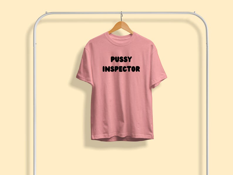 Pu&ograve; includere: Una maglietta rosa chiaro con la scritta "PUSSY INSPECTOR" stampata in grassetto nero. La maglietta &egrave; appesa a una gruccia di legno sospesa a un appendiabiti di metallo bianco su uno sfondo giallo chiaro.