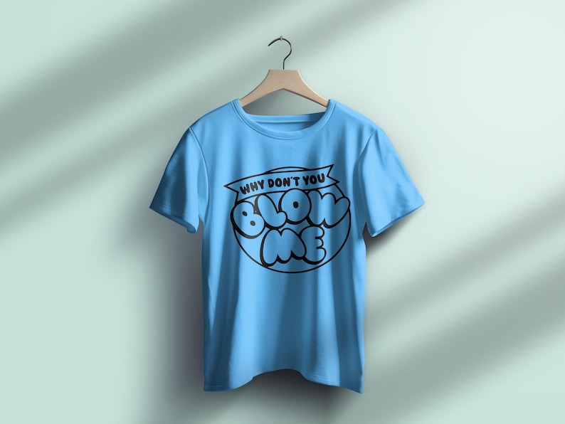 Pu&ograve; includere: T-shirt azzurra con la scritta nera "WHY DON'T YOU BLOW ME" in un cerchio. La maglietta &egrave; appesa a una gruccia di legno su uno sfondo azzurro chiaro.