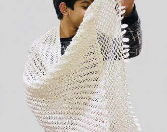 May include: A white, crocheted shawl with a triangular shape. The shawl is held up, showcasing the open weave and textured pattern. The edges are finished with a decorative trim. The shawl appears soft and lightweight.