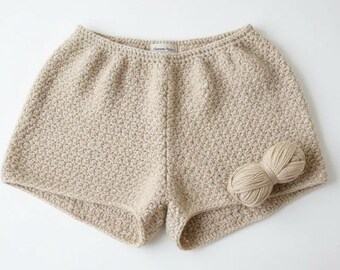 May include: Beige crochet shorts with a textured stitch pattern. A small ball of matching yarn rests beside the shorts. The shorts have a comfortable, relaxed fit, perfect for warm weather. The shorts are handmade.