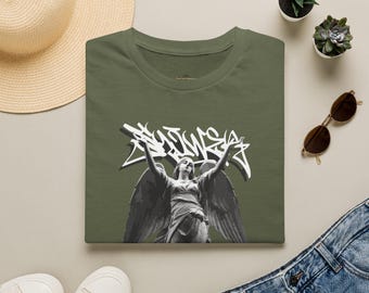 May include: An olive green t-shirt featuring a black and white graphic of a winged angel statue with arms raised. Above the angel is white graffiti-style text. The shirt has a crew neckline and short sleeves, presented in a flat lay composition.