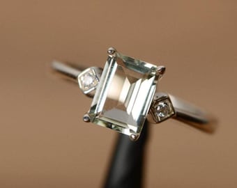Op de afbeelding: Zilveren ring met een rechthoekige, lichtgroene edelsteen als centraal element. Twee kleine, vierkante accentstenen met heldere facetten flankeren de hoofdsteen. De ring is geplaatst tegen een neutrale achtergrond.