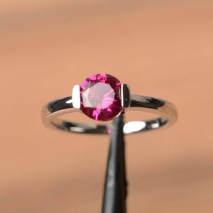 Könnte beinhalten: Ein silberner Ring mit einem runden, leuchtend rosa Edelstein. Der Edelstein ist in einem schlichten, eleganten Band gefasst. Der Ring wird vor einem neutralen Hintergrund gezeigt, der seine polierte Oberfläche und die Brillanz des Steins hervorhebt.
