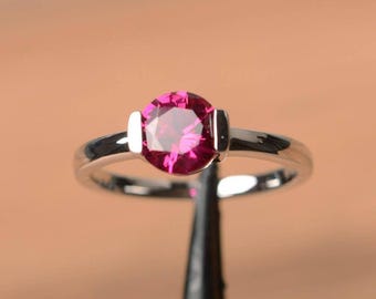 May include: A silver ring with a round, bright pink gemstone. The gemstone is set in a simple, elegant band. The ring is shown against a neutral background, highlighting its polished finish and the brilliance of the stone.