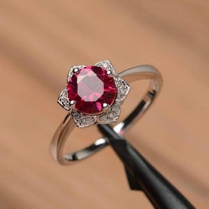 May include: A silver ring with a central, deep red gemstone, encircled by smaller, clear stones arranged in a floral design. The band is slender and polished. The ring's setting is flower-shaped. The background is blurred, focusing attention on the jewellery.
