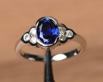 May include: A silver ring featuring a large, oval-cut, deep blue gemstone as the centerpiece. The ring is accented with smaller, round, clear gemstones on each side of the main stone. The setting has a decorative, floral-like design.