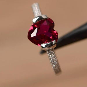 May include: A silver ring with a heart-shaped, deep red gemstone. The band is set with small, clear stones, and the heart is held by two silver clasps. The ring is shown against a blurred background.