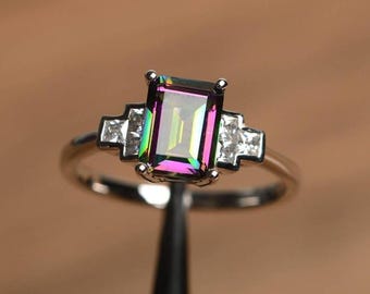 May include: A silver ring featuring a large, rectangular, iridescent gemstone with a rainbow effect. The ring has smaller, square, clear gemstones on each side of the main stone, set on a thin silver band.
