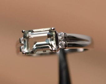 Op de afbeelding: Een zilveren ring met een rechthoekige, lichtgroene edelsteen als middelpunt. Kleine, heldere accentstenen zijn aan weerszijden van de hoofdsteen en op de band gezet. De ring heeft een klassiek, elegant ontwerp.