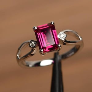 May include: A silver ring featuring a large, rectangular, vibrant pink gemstone as the centerpiece. Two smaller, square-cut clear stones flank the main stone. The ring's band has a unique, open design.