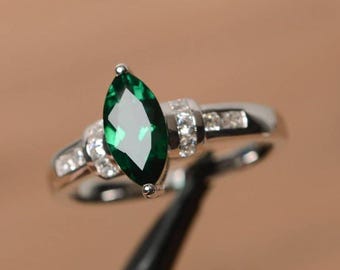 May include: A silver ring featuring a marquise-cut, emerald-green gemstone as the focal point. The band is adorned with smaller, clear gemstones. The ring is set against a blurred background.
