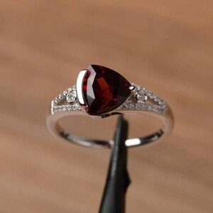 Puede incluir: Un anillo de tono plateado con una gran piedra preciosa granate de color rojo intenso y talla trillante como pieza central. El aro está dividido y adornado con pequeñas piedras de acento redondas y transparentes a lo largo de los hombros.