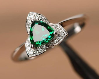 May include: A silver ring featuring a triangular, emerald-green gemstone at the center, surrounded by a floral-shaped setting of small, clear gemstones. The ring's band is thin and polished.