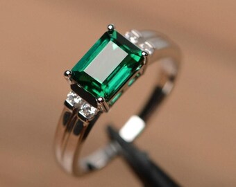 May include: A silver ring featuring a rectangular, emerald-cut green gemstone as the primary focal point. The ring has a split shank design and small accent stones on either side of the main stone. The ring is set against a blurred background.
