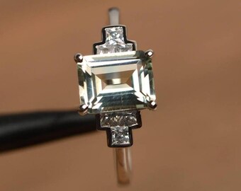 Op de afbeelding: Een zilveren ring met een grote, lichtgroene smaragdgeslepen edelsteen als middelpunt. De ring is versierd met kleinere, vierkant geslepen, heldere edelstenen aan weerszijden van de hoofdsteen, in een geometrisch ontwerp. De band is slank en gepolijst.