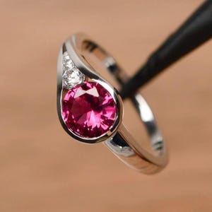Op de afbeelding: Zilveren ring met een grote, ronde, dieproze edelsteen. De ring heeft een split-schachtontwerp met drie kleinere, heldere edelstenen aan één kant van de hoofdsteen. De ring staat tegen een vage achtergrond.