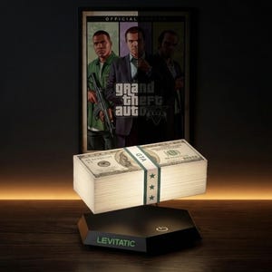May include: A stack of US $100 bills, secured with a white band and green stars, levitates above a black base. The base reads "LEVITATIC". A poster for "Grand Theft Auto V" is in the background.