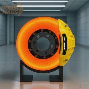 Op de afbeelding: Een oranje en zwarte remschijf met een gele McLaren remklauw, gemonteerd op een zwarte standaard. De schijf gloeit oranje. Het woord "McLaren" staat op de remklauw.