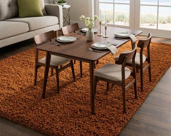 May include: A rectangular dark wood dining table with four chairs featuring light beige upholstered seats. The table is set with white plates, silverware, and a white vase with white flowers. It sits on a large, textured, burnt orange rug.