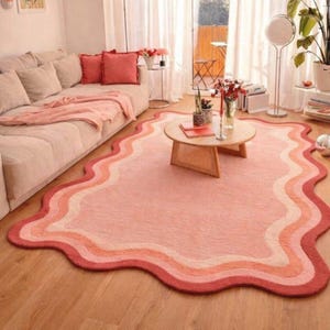 May include: A wavy-edged area rug in shades of pink, peach, and red. The rug features a layered design with a dark red border, transitioning through lighter shades to a pale pink center. The rug is placed on a wooden floor in a living room setting.