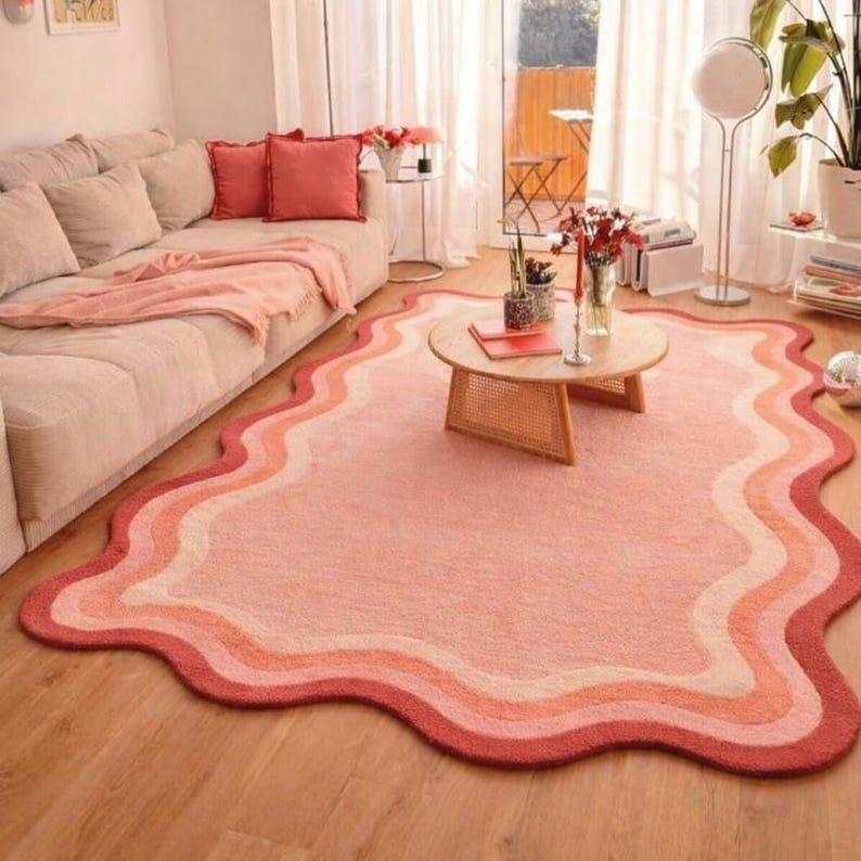 May include: A wavy-edged area rug in shades of pink, peach, and red. The rug features a layered design with a dark red border, transitioning through lighter shades to a pale pink center. The rug is placed on a wooden floor in a living room setting.