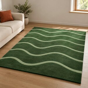 Puede incluir: Una alfombra rectangular que presenta un color base verde bosque profundo con líneas onduladas horizontales repetitivas en un tono verde salvia más claro. La alfombra tiene una superficie de pelo texturizado y está colocada sobre un suelo de madera.