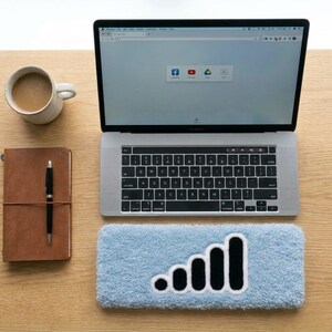 May include: A silver laptop with a black keyboard, a brown leather-bound notebook with a black pen, a white ceramic mug filled with coffee, and a light blue rectangular fuzzy wrist rest featuring a black signal strength icon.