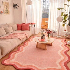 May include: A living room with a pink and peach color scheme. A wavy-edged rug in shades of pink and peach sits on a wooden floor. A beige sofa with coral pillows and a round coffee table complete the scene.