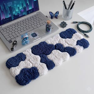 Puede incluir: Un reposamuñecas rectangular para teclado con una textura de pelo largo y mechones, que presenta un patrón abstracto e entrelazado de formas en azul marino profundo y blanco nítido. La superficie suave y afelpada ofrece apoyo al escribir.