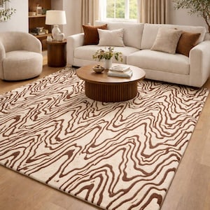 May include: A cream and brown patterned rug in a living room setting. The rug features a flowing, abstract design. The room also includes a white sofa, a round coffee table, and a comfortable armchair.