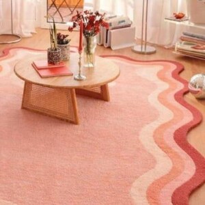 May include: A living room scene featuring a round wooden coffee table with a woven base, a wavy-edged rug in shades of pink and red, and decorative items. The rug's design adds a unique touch to the room's decor.