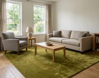 May include: A large, solid olive green area rug covering a light-toned wooden floor in a living room. The room features a beige sofa with two matching throw pillows, a light gray armchair, a wooden coffee table, and a side table with a lamp.