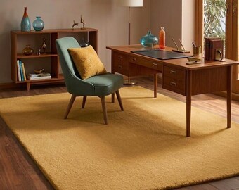 May include: A large rectangular mustard yellow area rug with a textured pile surface. The rug is positioned on a wooden floor in a room containing a wooden desk, a teal upholstered chair with a mustard yellow pillow, and a wooden bookshelf.