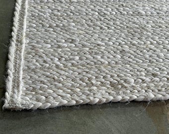 Puede incluir: Primer plano de una alfombra trenzada gris claro. La alfombra tiene un tejido grueso y texturizado con un borde trenzado. El color neutro y la textura natural sugieren una estética de diseño rústico o costero.