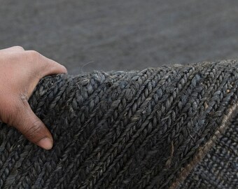 May include: Close-up of a dark grey woven rug. The rug has a thick, textured appearance with a braided pattern. The edge of the rug is visible, showing the thickness and construction. The rug is being held up, revealing its underside.