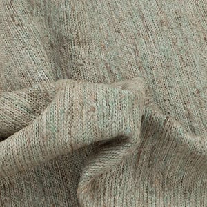 以下が含まれることがあります： ベージュと淡い緑色の織り目加工の生地のクローズアップ。生地は斑点模様で、柔らかく折りたたまれたドレープがあります。織り目がはっきりと見え、触覚的な表面を作り出しています。