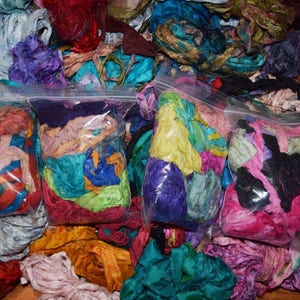 May include: Four bags of colorful silk ribbon scraps. The bags are filled with various shades of blue, green, pink, purple, and yellow ribbon scraps. The ribbon scraps are arranged in a random pattern.