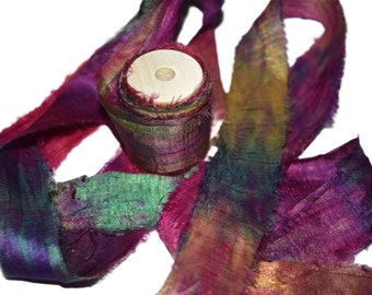 May include: A spool of colourful silk ribbon with a wooden centre. The ribbon is a mix of purple, green, gold, and red.