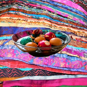 May include: A clear glass bowl filled with water and several decorated Easter eggs. The eggs are various colors, including red, green, and brown. The bowl sits on a colorful patchwork textile with vibrant stripes of pink, blue, orange, and purple.