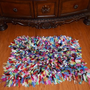 May include: A colorful, multi-colored, shaggy rug made from recycled materials. The rug is a rectangular shape and has a variety of colors, including pink, blue, green, red, and purple.